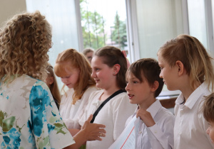 Zdjęcie przedstawia uczniów klasy 4a stojących w szeregu w sali gimnastycznej podczas uroczystości zakończenia roku szkolnego. Uczniowie ubrani są odświętnie – w białych bluzkach, koszulach, spódniczkach i spodenkach. Po lewej stronie stoi nauczycielka – wychowawczyni klasy, pani Paulina Klemżyńska, ubrana w jasne spodnie i kwiecistą bluzkę. Trzyma w rękach świadectwa, które wręcza kolejnym uczniom. W tle widać otwarte okna, papierowe torby z upominkami oraz inne osoby obserwujące wydarzenie.