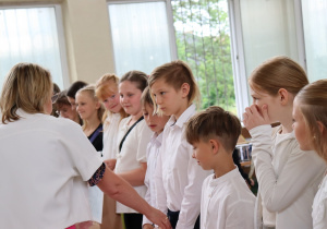 Zdjęcie przedstawia uczniów klasy 4b ustawionych w szeregu, odbierających świadectwa od swojej wychowawczyni, pani Małgorzaty Krawczyk, ubranej w białą marynarkę. Uczniowie mają na sobie galowe stroje: białe koszule, bluzki, spódnice. W tle widać okna sali gimnastycznej oraz zieleń za budynkiem. Dzieci wyglądają na skupione i przejęte chwilą, niektóre uśmiechają się do nauczycielki. Atmosfera zdjęcia jest uroczysta i podniosła.