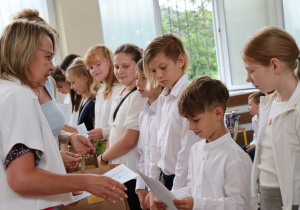 Zdjęcie przedstawia uczniów klasy 4b ustawionych w szeregu, odbierających świadectwa od swojej wychowawczyni, pani Małgorzaty Krawczyk, ubranej w białą marynarkę. Uczniowie mają na sobie galowe stroje: białe koszule, bluzki, spódnice. W tle widać okna sali gimnastycznej oraz zieleń za budynkiem. Dzieci wyglądają na skupione i przejęte chwilą, niektóre uśmiechają się do nauczycielki. Atmosfera zdjęcia jest uroczysta i podniosła.