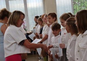 Zdjęcie przedstawia uczniów klasy 4b ustawionych w szeregu, odbierających świadectwa od swojej wychowawczyni, pani Małgorzaty Krawczyk, ubranej w białą marynarkę. Uczniowie mają na sobie galowe stroje: białe koszule, bluzki, spódnice. W tle widać okna sali gimnastycznej oraz zieleń za budynkiem. Dzieci wyglądają na skupione i przejęte chwilą, niektóre uśmiechają się do nauczycielki. Atmosfera zdjęcia jest uroczysta i podniosła.