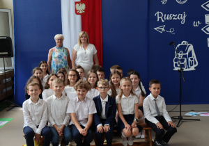 Zdjęcie przedstawia grupę uczniów klasy 4a Szkoły Podstawowej nr 13 w Pabianicach, ubranych w stroje galowe – białe koszule i ciemne spodnie lub spódniczki – siedzących i stojących w sali gimnastycznej na tle niebieskiej dekoracji z hasłem „Ruszaj w swoją podróż”. W tle stoją dwie nauczycielki: wychowawczyni klasy. Po lewej stronie widoczna jest biało-czerwona flaga z godłem Polski. Dzieci uśmiechają się i pozują do pamiątkowego zdjęcia.