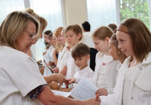 Zdjęcie przedstawia uczniów klasy 4b ustawionych w szeregu, odbierających świadectwa od swojej wychowawczyni, pani Małgorzaty Krawczyk, ubranej w białą marynarkę. Uczniowie mają na sobie galowe stroje: białe koszule, bluzki, spódnice. W tle widać okna sali gimnastycznej oraz zieleń za budynkiem. Dzieci wyglądają na skupione i przejęte chwilą, niektóre uśmiechają się do nauczycielki. Atmosfera zdjęcia jest uroczysta i podniosła.