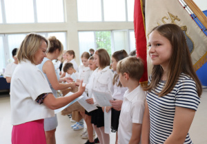 Zdjęcie przedstawia uczniów klasy 4b ustawionych w szeregu, odbierających świadectwa od swojej wychowawczyni, pani Małgorzaty Krawczyk, ubranej w białą marynarkę. Uczniowie mają na sobie galowe stroje: białe koszule, bluzki, spódnice. W tle widać okna sali gimnastycznej oraz zieleń za budynkiem. Dzieci wyglądają na skupione i przejęte chwilą, niektóre uśmiechają się do nauczycielki. Atmosfera zdjęcia jest uroczysta i podniosła.