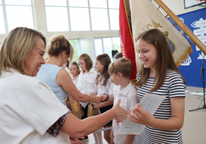 Zdjęcie przedstawia uczniów klasy 4b ustawionych w szeregu, odbierających świadectwa od swojej wychowawczyni, pani Małgorzaty Krawczyk, ubranej w białą marynarkę. Uczniowie mają na sobie galowe stroje: białe koszule, bluzki, spódnice. W tle widać okna sali gimnastycznej oraz zieleń za budynkiem. Dzieci wyglądają na skupione i przejęte chwilą, niektóre uśmiechają się do nauczycielki. Atmosfera zdjęcia jest uroczysta i podniosła.