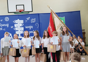 Zdjęcie przedstawia grupę wyróżnionych uczniów klasy 4b stojących na tle niebieskiej dekoracji z hasłem „Ruszaj w swoją podróż” i drogowskazem z napisem: marzenia, pasja, wiedza, przygoda. Uczniowie ubrani są w galowe stroje – białe koszule i bluzki, ciemne spódniczki lub spodnie – i trzymają w rękach świadectwa oraz papierowe torby z nagrodami. W tle widoczny jest poczet sztandarowy z czerwono-kremowym sztandarem szkoły oraz herbem Szkoły Podstawowej nr 13 w Pabianicach. W prawym dolnym rogu częściowo widoczna jest grupa siedzących uczniów.