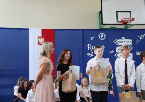 Zdjęcie przedstawia troje uczniów stojących w rzędzie w sali gimnastycznej Szkoły Podstawowej nr 13 w Pabianicach. Uczniowie ubrani są odświętnie i trzymają w rękach świadectwa, papierowe torby z nagrodami oraz kwiaty. Po lewej stronie stoi wychowawczyni, pani Renata Goszcz, ubrana w jasnoróżową sukienkę z tiulowym wykończeniem, uśmiechnięta i zwrócona w stronę uczniów. W tle znajduje się dekoracja z hasłem „Ruszaj w swoją podróż”, godło Polski, kosz do koszykówki oraz siedzący uczniowie. Atmosfera jest uroczysta i pełna radości.