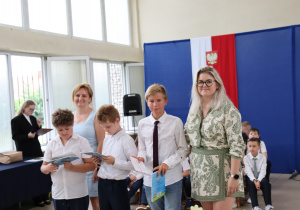 Zdjęcie przedstawia wyróżnionych uczniów klasy 5a stojących w rzędzie w sali gimnastycznej Szkoły Podstawowej nr 13 w Pabianicach. Uczniowie ubrani są odświętnie, trzymają świadectwa i torby z nagrodami. Po lewej stronie stoi dyrektor szkoły, pani Anna Michalak, ubrana w jasnoniebieską sukienkę. Po prawej stronie zdjęcia znajduje się wychowawczyni klasy 5a, pani Monika Kubiaczyk, ubrana w zieloną sukienkę z białym tłem i ornamentowym wzorem. W tle widoczna jest niebieska dekoracja z hasłem „Ruszaj w swoją podróż” oraz herbem szkoły. W głębi sali siedzą inni uczniowie.