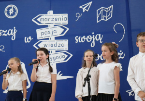 Zdjęcie przedstawia sześcioro uczniów klas trzecich występujących na scenie podczas uroczystości zakończenia roku szkolnego w Szkole Podstawowej nr 13 w Pabianicach. Troje dzieci trzyma mikrofony i śpiewa, pozostali recytują lub oczekują na swoją kolej. Dzieci ubrane są odświętnie – w białe koszule i ciemne spódnice lub spodnie. W tle widoczna jest niebieska dekoracja z białymi napisami „Ruszaj w swoją podróż” oraz drogowskazem z hasłami: marzenia, pasja, wiedza, przygoda. Dekorację uzupełniają rysunki papierowych samolotów i mapy.