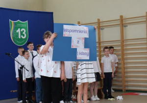 Zdjęcie przedstawia dwoje uczniów trzymających duży niebieski plakat z napisem „Wspomnienia z 1 klasy” na tle sali gimnastycznej Szkoły Podstawowej nr 13 w Pabianicach. Napis wykonany jest z trzech elementów ozdobionych kwiatowym motywem. W tle widoczni są pozostali uczniowie ubrani odświętnie w białe koszule i ciemne spodnie lub spódniczki. Po lewej stronie znajduje się mikrofon na statywie, a w tle herb szkoły oraz ściana gimnastyczna z drabinkami. Na podłodze leżą ozdobne papierowe kwiaty.