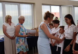 Zdjęcie przedstawia moment wręczania nagród uczniom klasy 4a. Po prawej stronie stoją uczniowie w galowych strojach – białych koszulach i ciemnych spodniach lub spódniczkach. Dyrektor szkoły, pani Anna Michalak, ubrana w niebieską sukienkę, ściska dłoń jednej z uczennic. Obok niej znajdują się nauczycielki: Violetta Kocik (w kwiecistej sukience bez rękawów) oraz Anna Wlazeł (w białej bluzce i jasnej, plisowanej spódnicy). W tle widać otwarte okna oraz stół z papierowymi torbami z nagrodami. Atmosfera jest oficjalna i uroczysta.