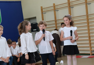 Zdjęcie przedstawia czworo uczniów klas trzecich biorących udział w części artystycznej podczas uroczystości zakończenia roku szkolnego. Dwoje dzieci trzyma mikrofony i mówi, pozostali przysłuchują się lub oczekują na swoją kolej. Wszyscy uczniowie są odświętnie ubrani – w białe koszule lub bluzki oraz ciemne spódnice lub spodnie. W tle widoczna jest sala gimnastyczna z drewnianymi drabinkami oraz siedzącymi uczniami. Na podłodze znajdują się dekoracyjne papierowe kwiaty.