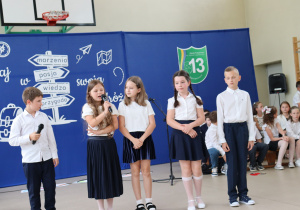 Zdjęcie przedstawia czworo uczniów klas trzecich biorących udział w części artystycznej podczas uroczystości zakończenia roku szkolnego. Dwoje dzieci trzyma mikrofony i mówi, pozostali przysłuchują się lub oczekują na swoją kolej. Wszyscy uczniowie są odświętnie ubrani – w białe koszule lub bluzki oraz ciemne spódnice lub spodnie. W tle widoczna jest sala gimnastyczna z drewnianymi drabinkami oraz siedzącymi uczniami. Na podłodze znajdują się dekoracyjne papierowe kwiaty.