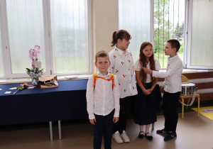 Zdjęcie przedstawia czworo uczniów klasy 3b stojących w sali gimnastycznej przy oknach. Troje dzieci rozmawia i uśmiecha się, jedno z nich (na pierwszym planie) pozuje do zdjęcia z niewielkim pomarańczowym plecakiem na ramionach. Dzieci ubrane są odświętnie w białe koszule, dziewczynka ma granatową spódnicę i długie, rozpuszczone włosy. Na stole w tle stoją kwiaty, papierowa torba i luźno położone upominki, obok widoczny jest bębenek na krześle. Atmosfera jest swobodna i radosna.