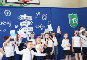 Zdjęcie przedstawia czworo uczniów klas trzecich biorących udział w części artystycznej podczas uroczystości zakończenia roku szkolnego. Dwoje dzieci trzyma mikrofony i mówi, pozostali przysłuchują się lub oczekują na swoją kolej. Wszyscy uczniowie są odświętnie ubrani – w białe koszule lub bluzki oraz ciemne spódnice lub spodnie. W tle widoczna jest sala gimnastyczna z drewnianymi drabinkami oraz siedzącymi uczniami. Na podłodze znajdują się dekoracyjne papierowe kwiaty.