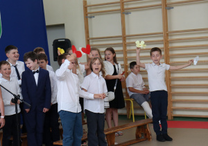 Zdjęcie przedstawia czworo uczniów klas trzecich biorących udział w części artystycznej podczas uroczystości zakończenia roku szkolnego. Dwoje dzieci trzyma mikrofony i mówi, pozostali przysłuchują się lub oczekują na swoją kolej. Wszyscy uczniowie są odświętnie ubrani – w białe koszule lub bluzki oraz ciemne spódnice lub spodnie. W tle widoczna jest sala gimnastyczna z drewnianymi drabinkami oraz siedzącymi uczniami. Na podłodze znajdują się dekoracyjne papierowe kwiaty.