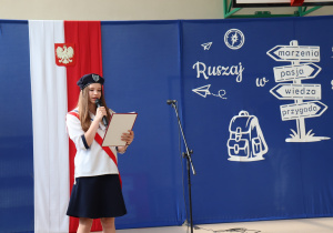 Zdjęcie przedstawia uczennicę stojącą na scenie sali gimnastycznej Szkoły Podstawowej nr 13 w Pabianicach. Ubrana jest w galowy strój pocztu sztandarowego – granatową spódnicę, białą bluzkę z granatowymi lamówkami i czerwono-biały szarf, a na głowie ma granatowy beret z numerem 13. Trzyma mikrofon i czyta z teczki. W tle widać dekorację: flagę Polski z godłem oraz niebieską planszę z białym napisem „Ruszaj w swoją podróż” i grafikami plecaka, drogowskazu oraz papierowych samolotów. Po prawej stronie widać mikrofon na statywie. Atmosfera jest oficjalna i podniosła.