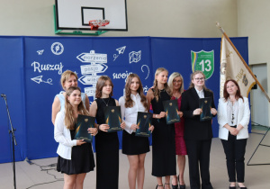 Zdjęcie przedstawia grupę wyróżnionych uczennic klasy 8a pozujących do pamiątkowego zdjęcia po wręczeniu świadectw ukończenia szkoły podstawowej. Uczennice ubrane są elegancko w czarne lub białe stroje i trzymają teczki z godłem Polski i napisem „Świadectwo ukończenia szkoły podstawowej”. Po lewej stronie stoi dyrektor szkoły, pani Anna Michalak, w jasnoniebieskiej sukience. Obok niej znajdują się wicedyrektor Edyta Makowska w bordowej sukience oraz wychowawczyni klasy Magdaleną Maciszewską w białej marynarce. W tle widoczna jest dekoracja z hasłem „Ruszaj w swoją podróż” oraz herbem szkoły i pocztem sztandarowym. Atmosfera jest uroczysta i pełna dumy.
