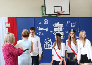 Zdjęcie przedstawia moment wręczenia nagród wyróżnionym uczniom klasy 8b. Po lewej stronie kadru widoczne są plecy dwóch kobiet – dyrektor szkoły Anny Michalak w jasnoniebieskiej sukience i wicedyrektor Edyty Makowskiej w różowej sukience z koronkowym wzorem. Na środku stoi uczeń w białej koszuli i okularach, odbierający gratulacje. Obok niego trzy uczennice – dwie ubrane w galowy strój pocztu sztandarowego z granatowymi beretami i biało-czerwonymi szarfami, a jedna w białej koszuli i czarnej spódnicy. W tle znajduje się dekoracja z drogowskazem i hasłem „Ruszaj w swoją podróż” oraz herbem szkoły. Atmosfera jest uroczysta.