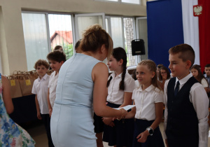 Zdjęcie przedstawia moment wręczania dyplomów uczniom klasy 3a przez dyrektor szkoły w jasnoniebieskiej sukience. Uczniowie ubrani są galowo – dziewczynki w białych bluzkach i granatowych spódniczkach, chłopiec w białej koszuli i ciemnej kamizelce. W tle widać innych uczniów oraz elementy dekoracji sali gimnastycznej.