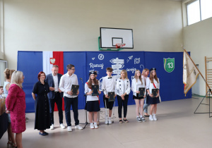 Zdjęcie przedstawia wyróżnionych uczniów klasy 8b stojących w szeregu wraz z rodzicami oraz nauczycielami na tle dekoracji sali gimnastycznej Szkoły Podstawowej nr 13 w Pabianicach. Uczniowie trzymają ciemne teczki ze świadectwami z wyróżnieniem. W tle znajduje się ścianka dekoracyjna z hasłem „Ruszaj w swoją podróż”, herbem szkoły oraz polską flagą i godłem. Po prawej stronie widoczny jest sztandar szkolny. Na twarzach uczestników widać dumę i radość z osiągnięć.