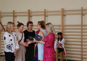 Zdjęcie przedstawia moment wręczania dyplomów podziękowania rodzicom uczniów klasy 8b przez dyrektor Annę Michalak oraz wychowawczynię Edytę Makowską. Po lewej stronie stoją rodzice w eleganckich strojach, z prawej strony w tle siedzi uczennica w stroju pocztu sztandarowego.