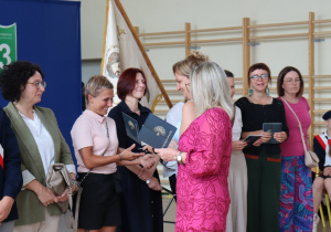 Zdjęcie przedstawia moment wręczania dyplomów podziękowania rodzicom uczniów klasy 8b przez dyrektor Annę Michalak oraz wychowawczynię Edytę Makowską. Po lewej stronie stoją rodzice w eleganckich strojach, z prawej strony w tle siedzi uczennica w stroju pocztu sztandarowego.