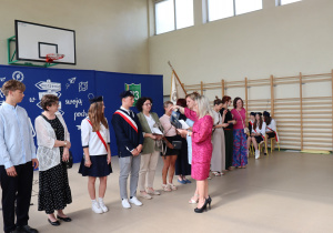 Zdjęcie przedstawia wręczanie dyplomów uznania przez dyrektor szkoły Annę Michalak i wicedyrektor Edytę Makowską rodzicom wyróżnionych uczniów klas ósmych. Po lewej stronie stoją również uczniowie klas ósmych, w tym członkowie pocztu sztandarowego. W tle widoczna jest ścianka dekoracyjna z napisem „Ruszaj w swoją podróż” oraz godło i sztandar szkoły.