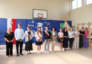 Zdjęcie przedstawia wręczanie dyplomów uznania przez dyrektor szkoły Annę Michalak i wicedyrektor Edytę Makowską rodzicom wyróżnionych uczniów klas ósmych. Po lewej stronie stoją również uczniowie klas ósmych, w tym członkowie pocztu sztandarowego. W tle widoczna jest ścianka dekoracyjna z napisem „Ruszaj w swoją podróż” oraz godło i sztandar szkoły.