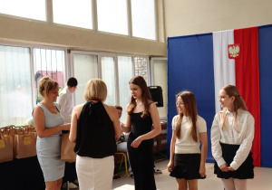 Na zdjęciu uczennice klasy 6a stojące w kolejce po odbiór nagród. Jedna z dziewcząt właśnie odbiera upominek od nauczycielki ubranej w czarną bluzkę i białą spódnicę – wychowawczyni Moniki Treichel. Obok stoi dyrektor szkoły Anna Michalak w jasnoniebieskiej sukience. W tle widać dekorację sali gimnastycznej z biało-czerwoną flagą i godłem Polski.