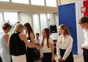 Na zdjęciu uczniowie klasy 6a podczas odbierania świadectw i nagród. Dziewczynka w okularach uśmiecha się i podaje rękę wychowawczyni Monice Treichel, ubranej w czarną bluzkę bez rękawów. Obok niej stoi dyrektor szkoły Anna Michalak w jasnoniebieskiej sukience. W tle biało-czerwona flaga z godłem Polski i dekoracja zakończenia roku szkolnego.