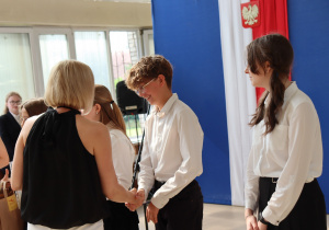 Na zdjęciu uczniowie klasy 6a w eleganckich strojach odbierają gratulacje od wychowawczyni Moniki Treichel, ubranej w czarną bluzkę bez rękawów i białą spódnicę. Chłopiec w okularach śmieje się i ściska dłoń nauczycielki. Obok niego stoją uczennice w białych koszulach. W tle dekoracja z flagą Polski i godłem.