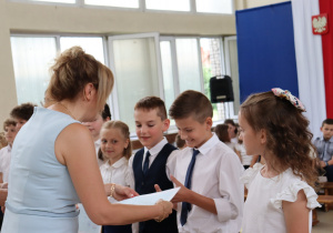 Na zdjęciu uczniowie klas 3a i 3b w galowych strojach odbierają świadectwa od dyrektor szkoły, ubranej w błękitną sukienkę. Dwaj chłopcy uśmiechają się, odbierając dokumenty. Obok stoją dziewczynki w białych bluzkach, a w tle widoczni są inni uczniowie i dekoracje z godłem Polski.