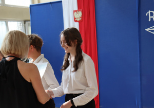 Na zdjęciu uczennice klasy 6a stojące w kolejce po odbiór nagród. Jedna z dziewcząt właśnie odbiera upominek od nauczycielki ubranej w czarną bluzkę i białą spódnicę – wychowawczyni Moniki Treichel. Obok stoi dyrektor szkoły Anna Michalak w jasnoniebieskiej sukience. W tle widać dekorację sali gimnastycznej z biało-czerwoną flagą i godłem Polski.