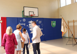 Na zdjęciu chłopiec z klasy 6b w białej koszuli odbiera świadectwo i prezent w torbie papierowej od dyrektor szkoły, ubranej w niebieską sukienkę. Obok stoją wicedyrektor w różowej sukience oraz wychowawca klasy 6b, Rafał Raźniewski, w jasnej marynarce w kratę. W tle niebieskie dekoracje z napisem „Ruszaj w swoją podróż”, logo szkoły oraz poczet sztandarowy.