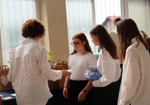 Na zdjęciu nauczycielka w białej koszuli, Sylwia Muszyńska, wręcza świadectwa uczennicom klasy 7a. Dziewczęta ubrane są w białe bluzki i czarne spódnice. Jedna z nich trzyma dwa niebieskie pudełka z upominkami. W tle widać stół z torbami prezentowymi oraz ozdobne storczyki.