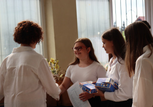 Nauczycielka w białej koszuli, widoczna tyłem, ściska dłoń uśmiechniętej uczennicy z klasy 7a, która trzyma świadectwo. Obok niej dwie koleżanki – jedna z nich trzyma dwa niebieskie pudełka z upominkami. W tle widać torby prezentowe i kwiaty ustawione na stole.