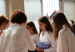 Na zdjęciu nauczycielka w białej koszuli, Sylwia Muszyńska, wręcza świadectwa uczennicom klasy 7a. Dziewczęta ubrane są w białe bluzki i czarne spódnice. Jedna z nich trzyma dwa niebieskie pudełka z upominkami. W tle widać stół z torbami prezentowymi oraz ozdobne storczyki.