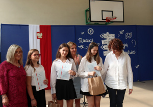 Pięć osób pozuje do wspólnego zdjęcia – trzy uczennice w białych koszulach trzymają świadectwa i torby z nagrodami, obok stoją dwie nauczycielki: jedna w bordowej sukience w kwiaty, druga w białej koszuli z rudymi kręconymi włosami. W tle niebieska dekoracja z hasłem „Ruszaj w swoją podróż” i symbolem Szkoły Podstawowej nr 13.