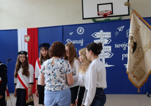 Nauczycielka w bluzce w kwiaty i niebieskich spodniach wręcza dokumenty uczennicom ubranym w białe koszule i czarne spódnice. W tle dekoracja z hasłem „Ruszaj w swoją podróż”, godło Polski oraz uczennice pocztu sztandarowego w mundurkach z czerwonymi szarfami.