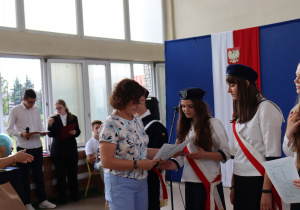 Grupa uczennic w białych bluzkach i czarnych spódnicach odbiera nagrody od nauczycielki w jasnej bluzce we wzory i niebieskich spodniach. W tle dekoracja z hasłem „Ruszaj w swoją podróż”, godło Polski oraz sztandar szkoły z napisem „Ojczyzna Nauka Praca”. Po lewej stronie stoją członkowie pocztu sztandarowego w mundurkach szkolnych z czerwonymi szarfami.