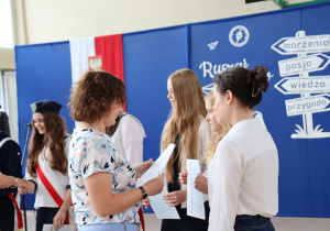 Nauczycielka w bluzce w kwiaty i niebieskich spodniach wręcza dokumenty uczennicom ubranym w białe koszule i czarne spódnice. W tle dekoracja z hasłem „Ruszaj w swoją podróż”, godło Polski oraz uczennice pocztu sztandarowego w mundurkach z czerwonymi szarfami.