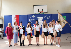 Grupa siedmiorga uczniów ubranych odświętnie, część w mundurach pocztu sztandarowego, pozuje do zdjęcia z dyplomami i torbami upominkowymi. Towarzyszą im trzy kobiety – nauczycielki i dyrekcja szkoły. W tle dekoracja z hasłem „Ruszaj w swoją podróż”, godło Polski oraz szkolny sztandar.