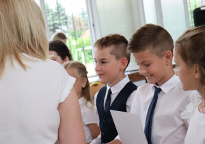 Grupa uczniów z klasy 3a ubranych odświętnie – chłopcy w białych koszulach i krawatach, dziewczynka z kokardą we włosach – uśmiecha się i spogląda na otrzymywane świadectwa. Na pierwszym planie nauczycielka w jasnej bluzce wręcza dokumenty. W tle widać salę z dużymi oknami i krzesłami.