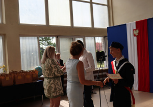 Chłopiec w granatowym stroju galowym z czerwono-białą szarfą i beretem, trzymający dyplom, stoi na tle dekoracji z godłem Polski oraz napisem „Ruszaj w swoją podróż”. Po lewej stronie kobiety wręczają uczniowi nagrodę. W tle widać torby upominkowe ustawione na stole oraz duże okna, przez które wpada światło dzienne.