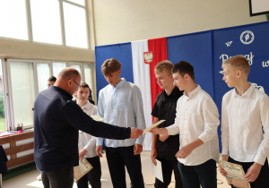 Grupa uczniów w eleganckich koszulach odbiera dyplomy od nauczyciela wychowania fizycznego, ubranego w granatową bluzę. Uczniowie stoją w szeregu, trzymając w rękach dyplomy. W tle widoczna dekoracja z hasłem „Ruszaj w swoją podróż” oraz biało-czerwona flaga z godłem Polski. Uroczystość odbywa się w jasnej sali gimnastycznej z dużymi oknami.