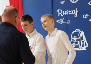 Grupa uczniów w eleganckich koszulach odbiera dyplomy od nauczyciela wychowania fizycznego, ubranego w granatową bluzę. Uczniowie stoją w szeregu, trzymając w rękach dyplomy. W tle widoczna dekoracja z hasłem „Ruszaj w swoją podróż” oraz biało-czerwona flaga z godłem Polski. Uroczystość odbywa się w jasnej sali gimnastycznej z dużymi oknami.