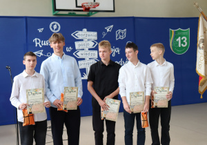 Pięciu uczniów w odświętnych strojach stoi na tle niebieskiej dekoracji z hasłami: „marzenia”, „pasja”, „wiedza”, „przygoda”. Każdy z nich trzyma w rękach dyplom sportowy oraz pomarańczowy worek z nagrodą. W tle widoczne godło Polski, tarcza Szkoły Podstawowej nr 13 w Pabianicach oraz sztandar. Zdjęcie wykonano w sali gimnastycznej podczas uroczystości szkolnej.