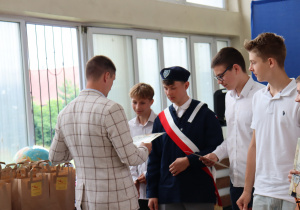 Nauczyciel w jasnej marynarce wręcza dyplomy grupie uczniów ubranych odświętnie. Jeden z chłopców ma strój pocztu sztandarowego z granatowym mundurem i beretem. W tle widoczne są okna sali gimnastycznej, torby z nagrodami oraz fragment niebieskiej dekoracji z emblematem Szkoły Podstawowej