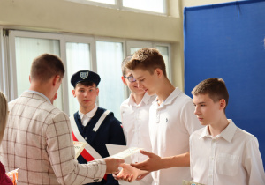 Nauczyciel w jasnej marynarce wręcza dyplomy grupie uczniów ubranych odświętnie. Jeden z chłopców ma strój pocztu sztandarowego z granatowym mundurem i beretem. W tle widoczne są okna sali gimnastycznej, torby z nagrodami oraz fragment niebieskiej dekoracji z emblematem Szkoły Podstawowej