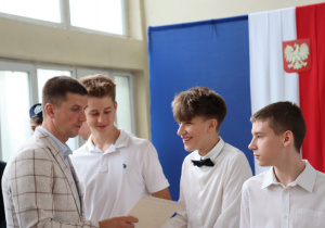 Nauczyciel w jasnej marynarce wręcza dyplomy grupie uczniów ubranych odświętnie. Jeden z chłopców ma strój pocztu sztandarowego z granatowym mundurem i beretem. W tle widoczne są okna sali gimnastycznej, torby z nagrodami oraz fragment niebieskiej dekoracji z emblematem Szkoły Podstawowej